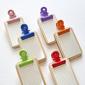 May include: Six small clipboards with colorful clips in pink, purple, blue, orange, green, and red. Each clipboard has a grid-lined paper and a natural wood-colored backing.