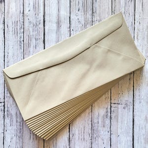 May include: A pile of beige envelopes, angled and neatly arranged, on a light-coloured, weathered wooden surface. The rectangular envelopes have a classic flap design, suitable for stationery or invitations.