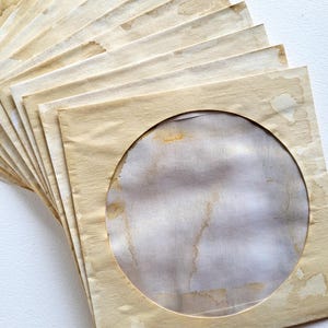 Könnte beinhalten: Ein Stapel gealterter Papierhüllen mit kreisförmigen Aussparungen, wahrscheinlich für Schallplatten oder Fotos. Das Papier hat einen warmen, beigen Farbton mit dunkleren braunen Flecken, was auf Alter oder eine Vintage-Ästhetik hindeutet. Die Hüllen sind fächerförmig angeordnet.