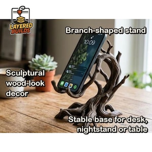 May include: A dark brown, branch-shaped phone stand with a textured, wood-like finish. The sculptural stand features a stable, wide base and curved prongs designed to hold a smartphone upright. The logo for Layered Builds is visible.