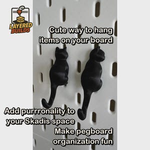 May include: Two black cat-shaped hooks attached to a white pegboard. The cats are depicted from behind with their tails curving downward to form hooks. Text on the image includes "LAYERED BUILDS", "Cute way to hang items on your board", "Add purrronality to your Skadis space", and "Make pegboard organization fun".