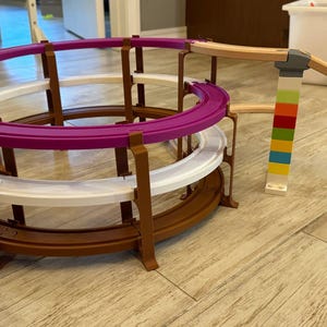 May include: A multi-level toy track set with a purple, white, and brown circular track. The track is supported by brown pillars. A wooden ramp connects to colorful block towers, creating a dynamic play structure.