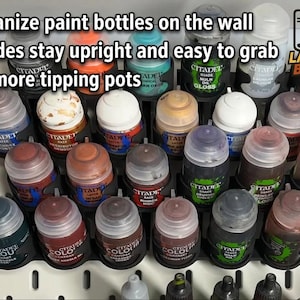 May include: A black plastic wall-mounted paint bottle organizer holding multiple Citadel paint pots in various colors. The organizer is attached to a white pegboard. Text on the image reads: Organize paint bottles on the wall, Shades stay upright and easy to grab, No more tipping pots, and LAYERED BUILDS.