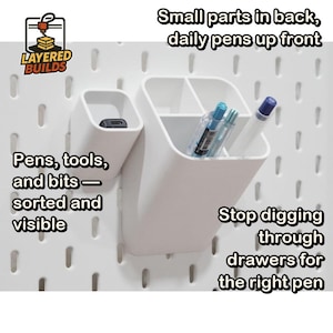 May include: A white plastic pegboard organizer system featuring two separate compartments mounted on a white pegboard. The larger container has three sections holding pens, while the smaller container holds a black tool bit. Text includes Layered Builds.