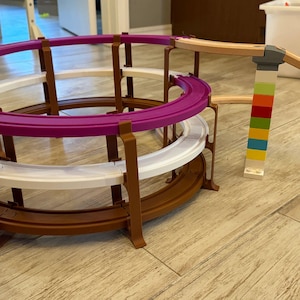 May include: A multi-level toy track set with a purple, white, and brown circular track. The track is supported by brown pillars. A wooden ramp connects to colorful block towers, creating a dynamic play structure.