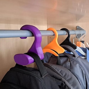 May include: Three S-shaped plastic hooks in purple, orange, and black hanging on a metal closet rod. Each hook features a wide, curved base designed to support the top handle of a backpack, keeping the bags organized and suspended in a row.