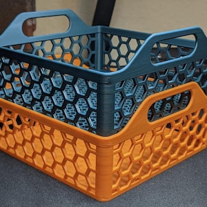 May include: Two plastic storage baskets, one teal and one orange, stacked on top of each other. The baskets have a honeycomb pattern and handles for easy carrying. The baskets are ideal for organizing items.
