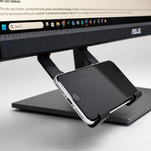 May include: A black plastic smartphone mount attached to the underside of an ASUS computer monitor. The mount holds a silver and black smartphone in a landscape orientation, positioned directly below the screen's bottom edge and taskbar.