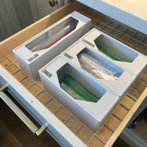 May include: A kitchen drawer with white organizers labeled "Gallon", "Quart", "Snack", and "Sandwich". Each organizer holds clear plastic bags with red, green, and clear contents. The drawer has a wooden interior and a metal grid.