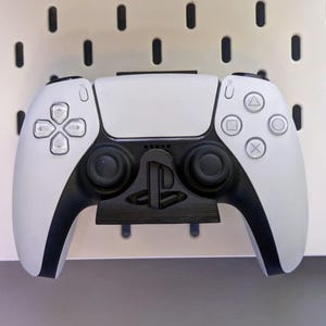 May include: A white and black game controller with a black stand, mounted on a white pegboard. The controller has a symmetrical design with buttons and joysticks. The pegboard has evenly spaced holes.