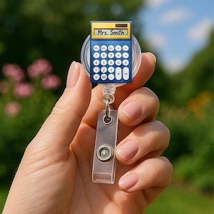May include: A blue calculator-shaped badge reel with the name "Mrs. Smith" displayed on the screen. The reel has a clear plastic clip and a retractable cord. The background is a blurred view of green foliage.