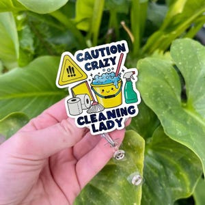 May include: A badge reel with a colorful cleaning-themed design. The badge features a yellow caution sign, a smiling yellow bucket with blue bubbles, a green spray bottle, and the text "CAUTION CRAZY CLEANING LADY".