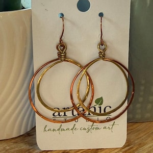 May include: A pair of dangle earrings featuring two interlocking circular hoops, one in a bright gold tone and one in a copper tone, suspended from wire hooks. The earrings are displayed on a white card with the text artenic studio handmade custom art.