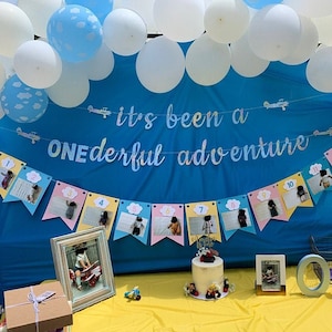 May include: A blue and white banner with the text "It's been a ONEderful adventure" hangs above a table with a cake and framed photos. The table is decorated with blue and white balloons.