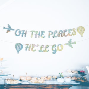 May include: A banner with the text "OH THE PLACES HE'LL GO" in blue letters, decorated with airplane and hot air balloon shapes. The letters and shapes are patterned with a map design. The banner is strung above a table with food.