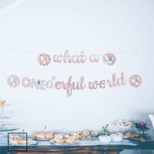 May include: A white banner with the text "what a wonderful world" in pink and white letters. The banner is decorated with small globe-shaped cutouts. The banner is hanging on a white wall above a table with food and drinks.