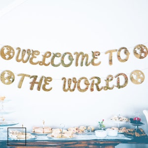 May include: Welcome to the world banner with a vintage map design. The banner is strung with twine and features the words "Welcome to the World" in gold letters with a world map design. The banner is decorated with small world map cutouts.