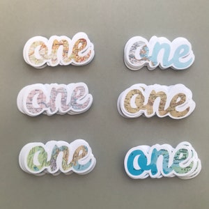 May include: A collection of die-cut stickers spelling the word "one". The stickers feature a white border and a map design in various colors, including blue, yellow, and green. The stickers are likely for crafts or party decorations.