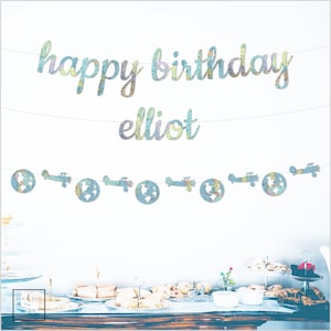 May include: A blue and white map-themed birthday banner with the words "happy birthday elliot" and a garland of paper airplanes and globes.