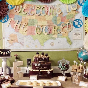 May include: A decorated table with desserts and a map backdrop. The map reads "Welcome to the World". Desserts include cakes, cookies, and candies. Decorations include paper fans, a globe, and wooden letters.