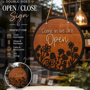 May include: Double-sided wooden open/close sign. The sign is round with a dark brown border and a lighter brown center. The text "Come in we are Open" is displayed on one side, and "Sorry we are Closed" on the other. The sign is decorated with floral designs and hangs by a jute rope.
