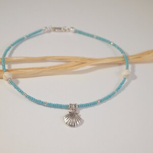 May include: Anklet with turquoise beads and silver accents. The beaded strand features a small seashell charm and two white, round beads. The anklet is secured with a silver clasp. A delicate piece of jewelry.