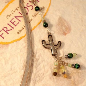 May include: A silver bookmark with a cactus charm and dangling green and yellow beads. The bookmark has a tag that reads "FRIENDSHIP" in purple letters.
