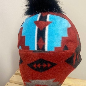 May include: A winter hat with a Southwestern-inspired design. The hat features a red, turquoise, and black geometric pattern. A fluffy black pom-pom sits atop the hat. The hat is made of a soft, warm material, perfect for cold weather.