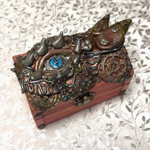 May include: A rectangular wooden box with a reddish-brown finish, featuring a decorative lid adorned with sculpted metallic elements, including a central blue eye, various gears, spikes, and swirling patterns, secured by a small metal latch.