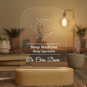Può includere: Una luce notturna personalizzata con base in legno e pannello in acrilico trasparente. Il pannello presenta un design a mezzaluna e stelle, insieme al testo "Sleep Medicine Sleep Specialist Dr. Chris Davis."