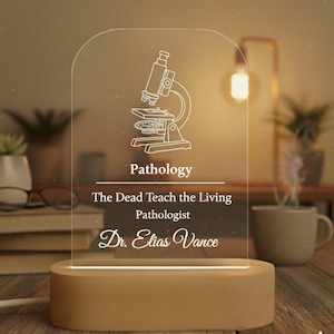 May include: A lit acrylic sign featuring a microscope illustration and the word "Pathology." The text below reads "The Dead Teach the Living Pathologist Dr. Elias Vance." It is set on a wooden base, giving off a warm light.