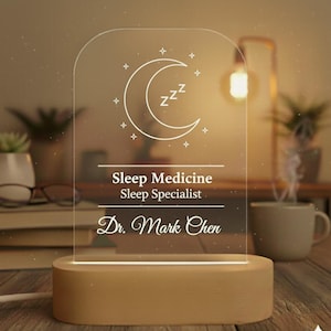 Può includere: Una luce notturna in acrilico trasparente con base in legno. La luce presenta un design a mezzaluna e stelle, con il testo "Sleep Medicine Sleep Specialist Dr. Mark Chen" inciso sul davanti. La luce è accesa, illuminando la stanza.