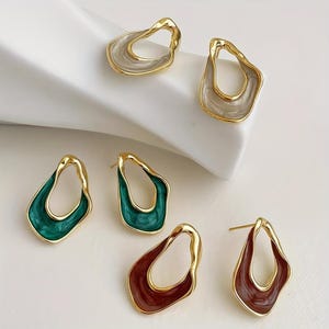 Peut inclure: Trois paires de boucles d'oreilles à tige avec des cadres en métal doré aux formes irrégulières et ouvertes. Chaque paire présente un émail de couleur différente : gris clair, vert émeraude profond et un brun rougeâtre foncé et riche.