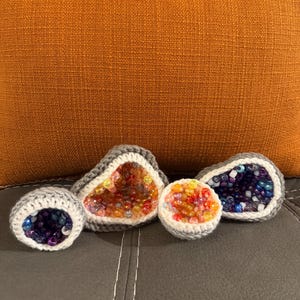 May include: Four crocheted geode sculptures with colorful bead interiors. The geodes are in shades of gray and white, with the bead colors including orange, yellow, blue, and purple. The geodes are displayed on a dark gray surface with an orange pillow in the background.