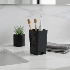May include: A black toothbrush holder with three toothbrushes and a small succulent plant in a black pot. A gray folded towel is next to the holder. The holder has a rectangular shape with cut-out designs.