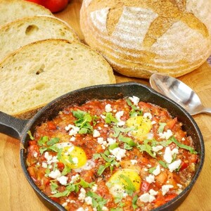 Puede incluir: Una sartén de hierro fundido negra que contiene shakshuka con tres huevos escalfados, una salsa a base de tomate, queso blanco desmenuzado y hierbas verdes, acompañada de una hogaza de pan crujiente y tres rebanadas.