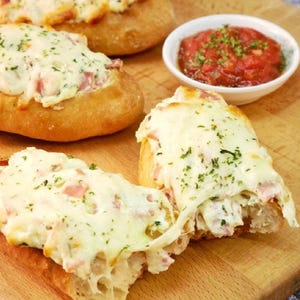 Puede incluir: Tres panecillos dorados cubiertos con queso blanco derretido, jamón en dados y hierbas verdes, acompañados por un pequeño cuenco blanco lleno de una salsa de tomate roja y espesa, todo dispuesto sobre una superficie de madera clara.