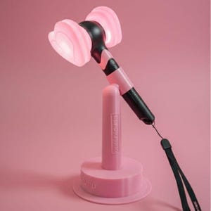 May include: A pink and black light stick with heart-shaped light heads, resting on a matching pink stand. The stand has the text "BLACKPINK" and "SUFU" printed on it. A black wrist strap is attached to the light stick.