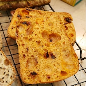 Puede incluir: Una rebanada de pan de color naranja dorado con una estructura de miga porosa y aireada con inclusiones oscuras visibles, sobre una rejilla de alambre negra. El pan presenta una corteza tostada y una textura interior suave y abierta.