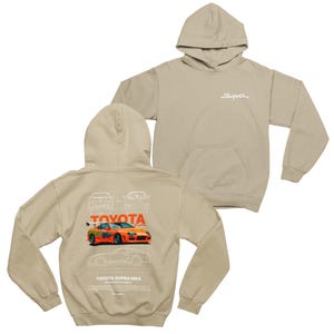 Könnte beinhalten: Zwei beige Kapuzenpullover. Einer hat das Wort "Supra" in Weiß auf der Brust. Der andere zeigt eine Grafik eines orangefarbenen Toyota Supra MK4 Autos auf der Rückseite, mit "Toyota" darüber. Die Grafik enthält auch Autoschemata.