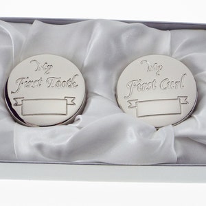 May include: Two silver keepsake boxes with engraved lids. The box on the left says "My First Tooth" and the box on the right says "My First Curl". Both boxes have a blank space for engraving a name.