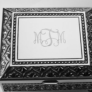 May include: A silver-coloured rectangular box with intricate engraved detailing. The lid has a central panel with stylized initials. The box has a decorative border with a repeating pattern and a small clasp on the front.