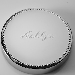 May include: A shiny, silver-colored, round lid with a beaded edge. The name "Ashlyn" is elegantly engraved in a cursive font on the lid's surface. The lid has a polished, reflective finish.