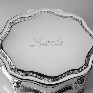 May include: A silver-toned jewelry box with a scalloped edge and ornate detailing. The lid is engraved with the name "Lucie" in cursive script. The box is elevated on decorative feet, suggesting a vintage or heirloom design.