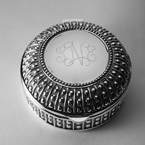 May include: A silver-colored, round trinket box with a hinged lid. The lid features an engraved monogram within a circular frame. The box has a detailed, textured design with vertical ribbed patterns and small, raised dots. The base has a decorative border.