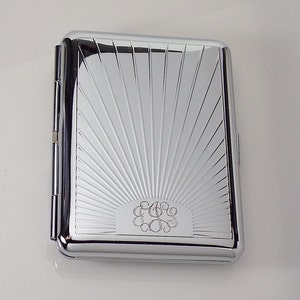 May include: A silver cigarette case with a sunburst design and a monogrammed initial in the center.