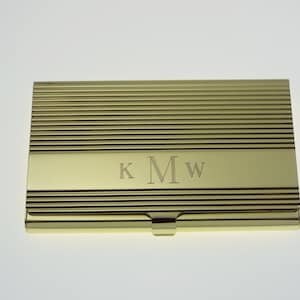 May include: A gold-colored business card holder with a horizontally ribbed design. The initials "KMW" are engraved on the front. The card holder has a rectangular shape and a small clasp.
