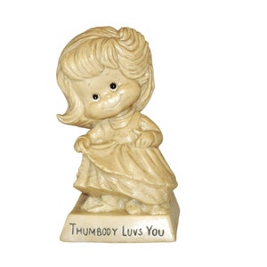May include: A vintage figurine of a smiling girl in a dress, holding her skirt. The figurine is a cream color with black eyes and the text "THUMBODY LUV'S YOU" on the base.