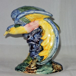 May include: A ceramic bird figurine with blue, green, yellow, and pink paint. The bird is perched on a tree branch with a brown, green, and blue base.