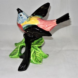 May include: A colorful ceramic bird figurine perched on a branch with green leaves. The bird has black, pink, yellow, blue, and green paint.